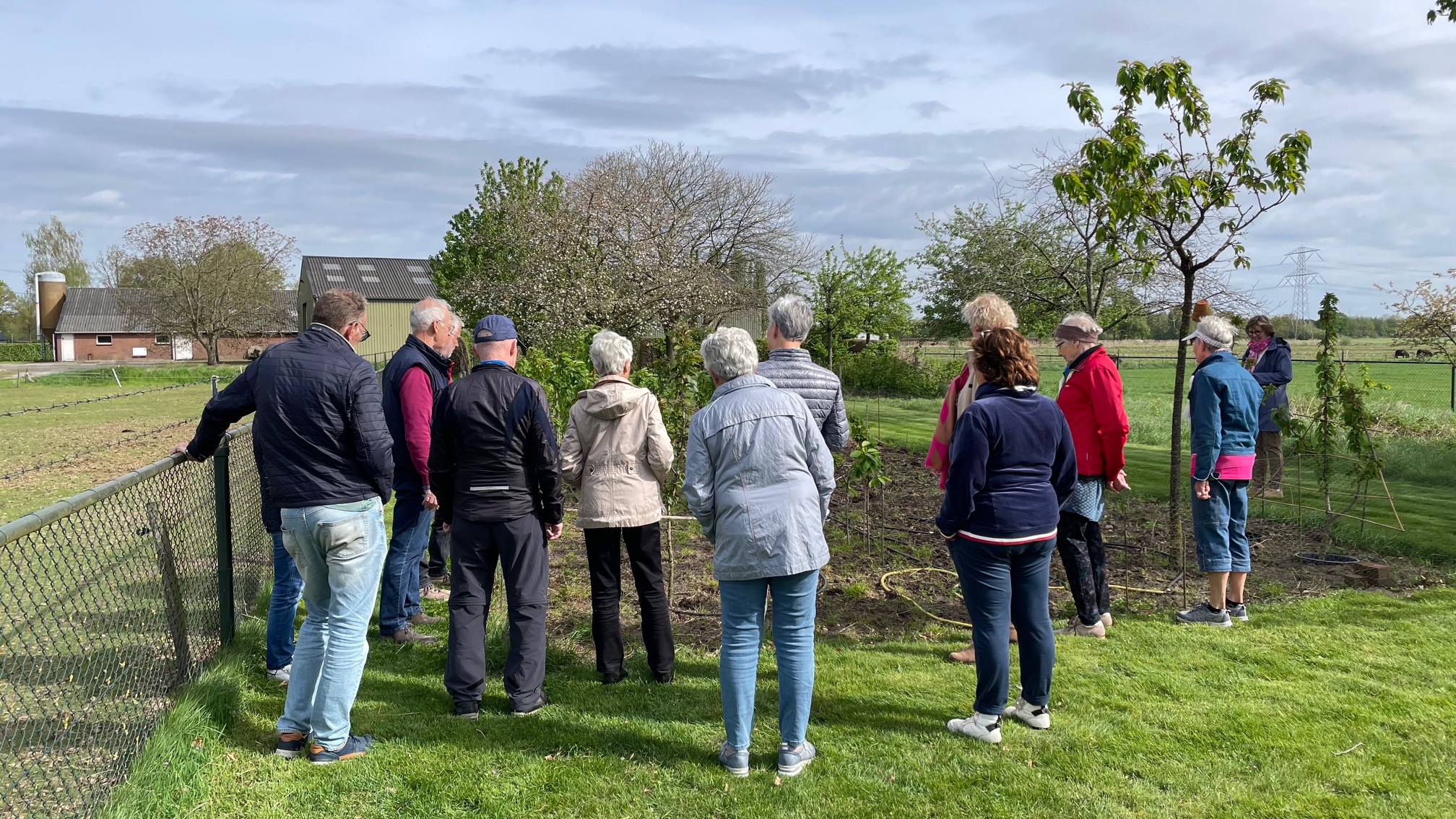 Een groep mensen staat buiten bij een tuin, met bomen en een hek op de achtergrond.