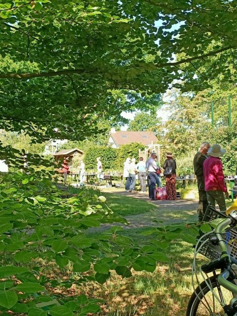 Mensen verzamelen zich in een groene tuin, met een fiets op de voorgrond.