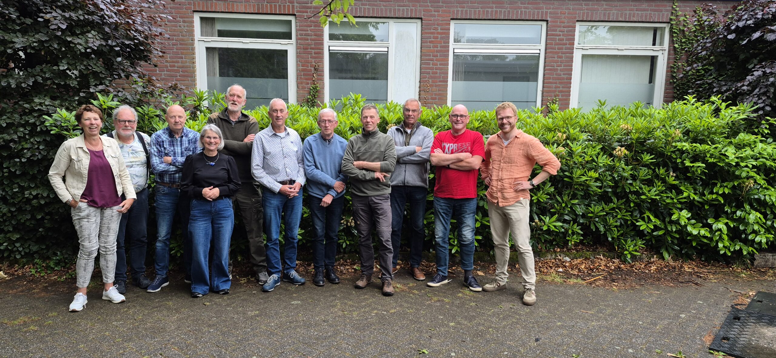 Groep mensen poseert buiten voor een gebouw met groene heg op achtergrond.