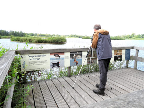 Man met telescoop kijkt naar vogels bij een meer, bij informatiebord "Vogeledorado de Kwegt".