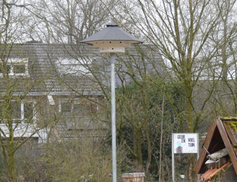 Een straatlantaarn met een grijze kap naast een bordje met "GEUR EN VOEL TUIN" en een huis op de achtergrond.