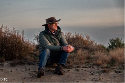 Man in groene jas en hoed zit in een zandlandschap bij zonsondergang, omringd door struikgewas.
