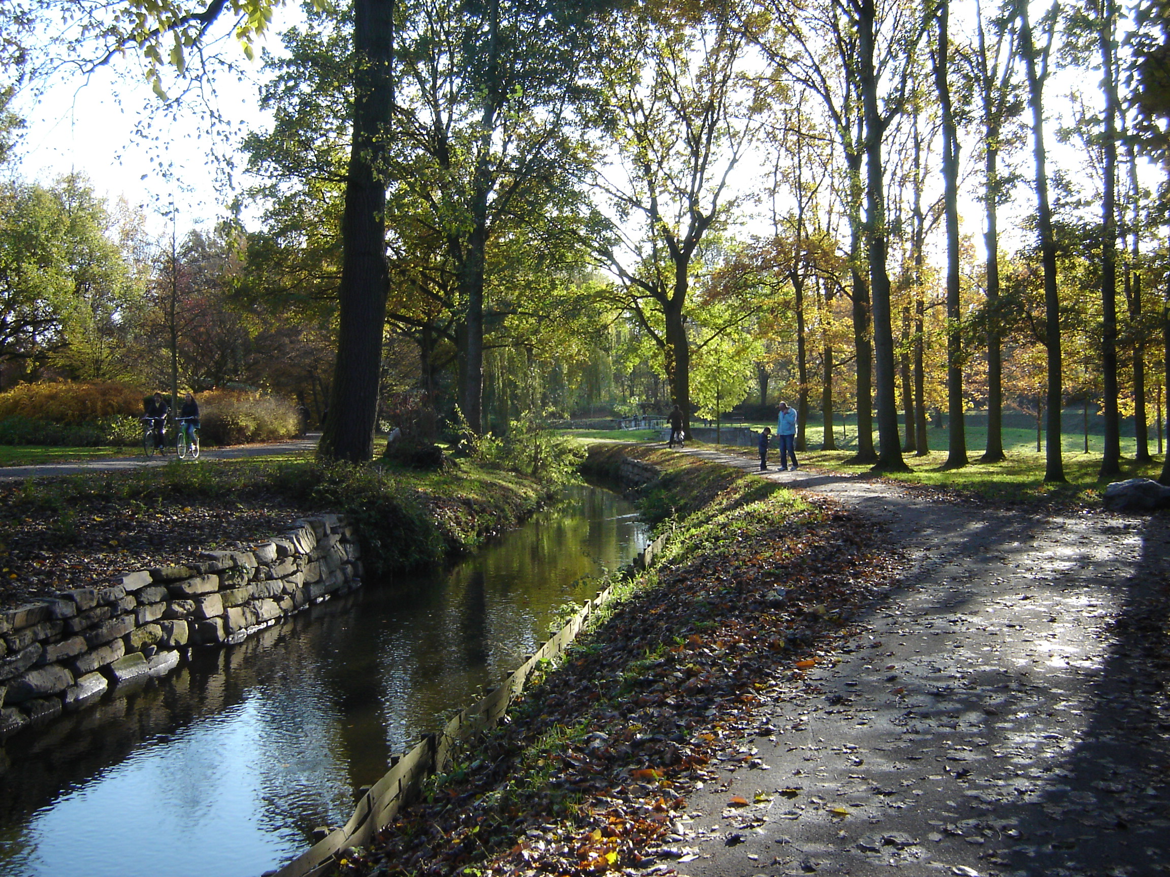 Bosrijk pad langs kabbelende beek, met wandelaars en fietsers in de herfstzon.