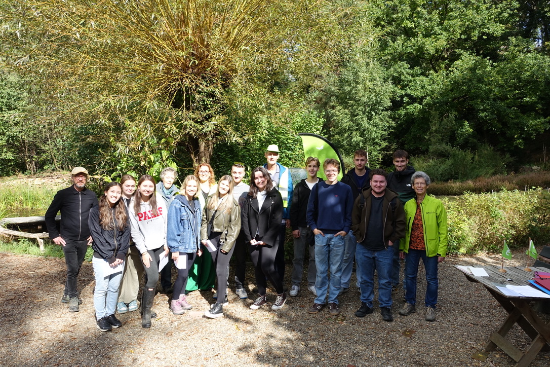 Een groep mensen poseert buiten bij een boom op een zonnige dag, omgeven door groen.