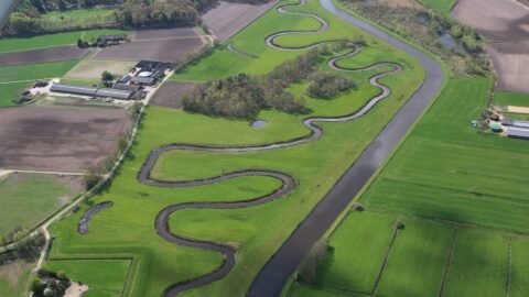 Luchtfoto van kronkelende rivier door groene velden met omliggende boerderijen en bossen.