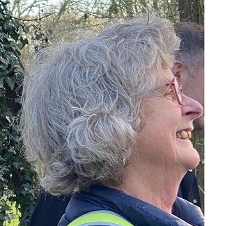 Een lachende vrouw met grijs haar draagt een bril, buiten in een groene omgeving.