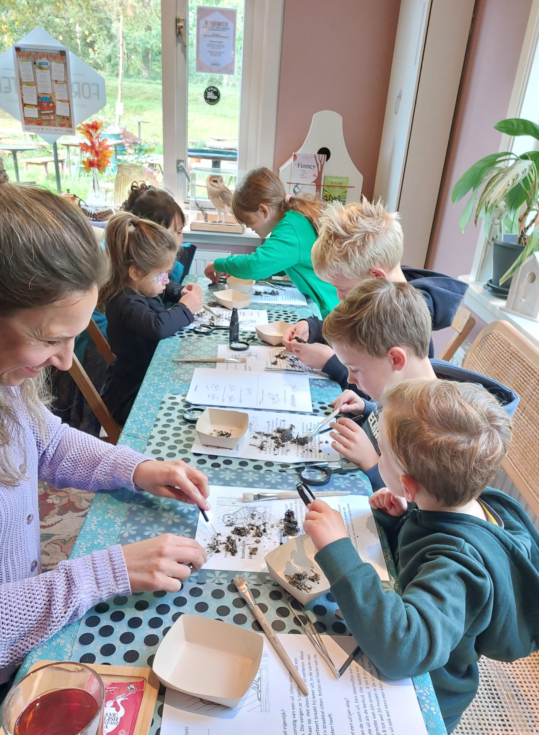 Kinderen en een volwassene doen een natuuronderzoek aan een tafel met loepen en papieren.