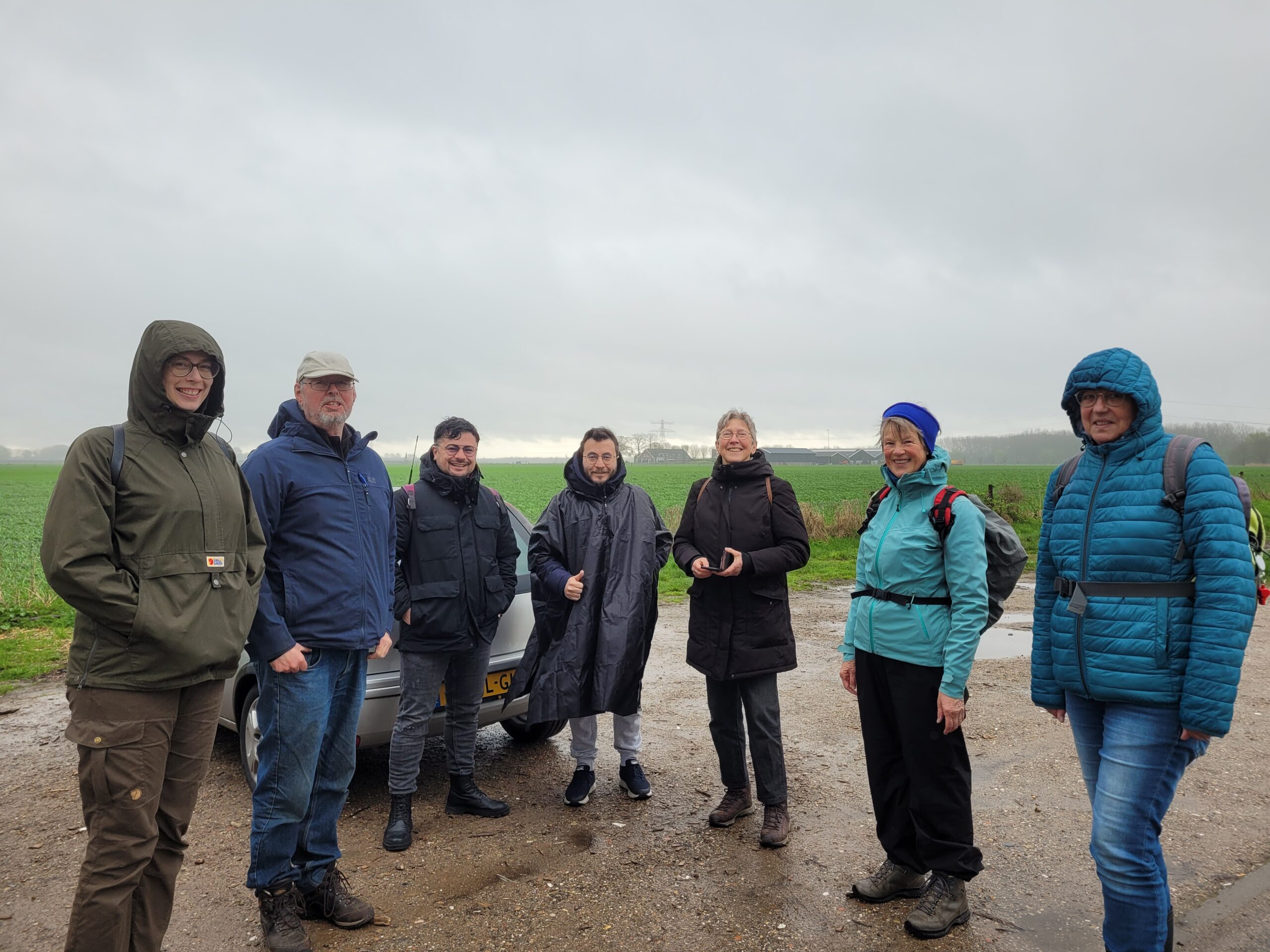 Groep van zeven mensen in buitenkleding op een regenachtige dag, staand op een onverharde weg.