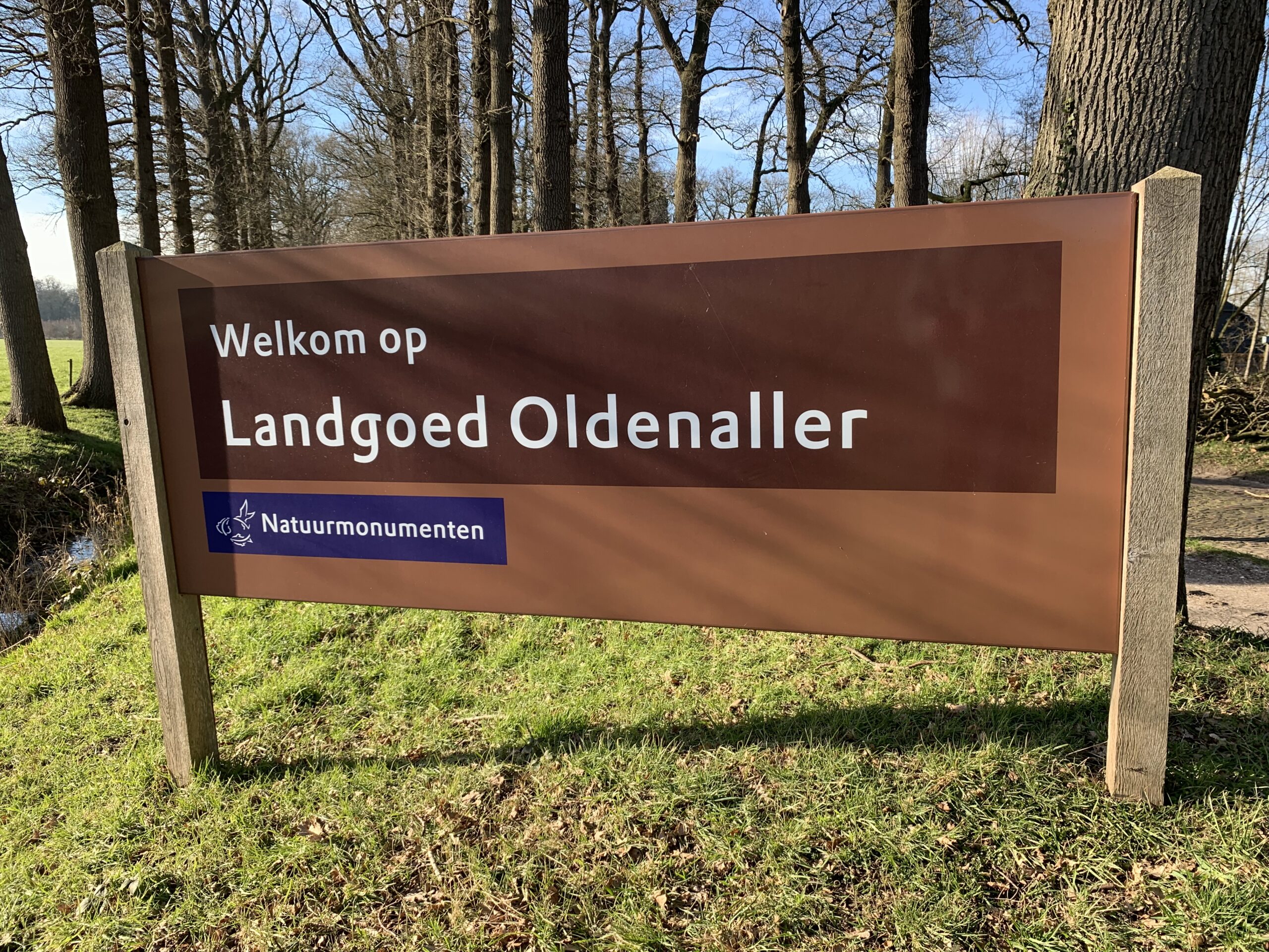 Bord met "Welkom op Landgoed Oldenaller" in bosrijke omgeving, logo Natuurmonumenten.