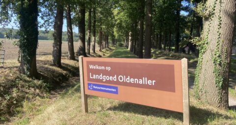 Welkomstbord bij Landgoed Oldenaller in een bosrijke omgeving met hoge bomen.