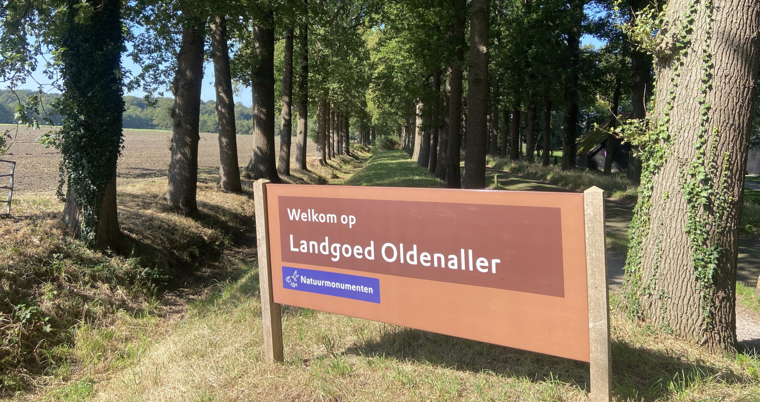 Welkomstbord bij Landgoed Oldenaller in een bosrijke omgeving met hoge bomen.