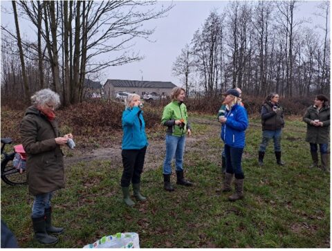 Groep mensen in buitenkleding drinkt koffie in een grasveld, omringd door bomen.