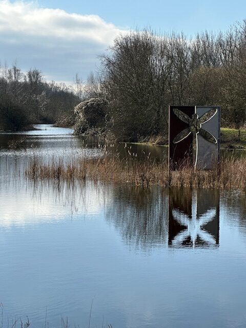 Een kunstwerk met libellemotief aan de oever van een rustige rivier onder een deels bewolkte hemel.
