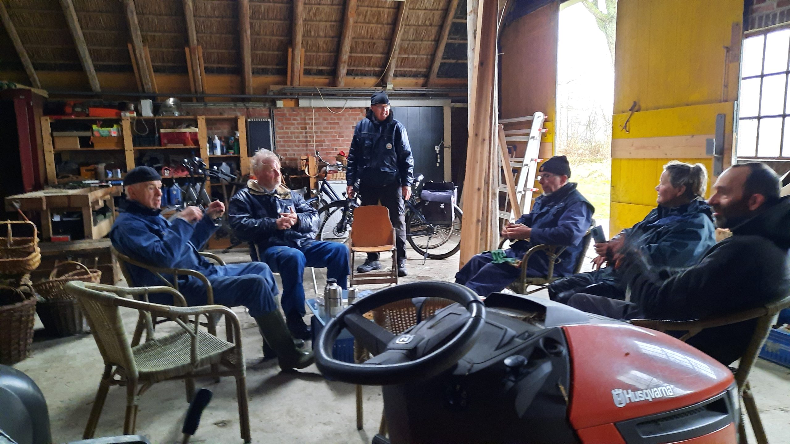 Groep mensen in een schuur, zittend rondom, met fietsen en gereedschap op de achtergrond.