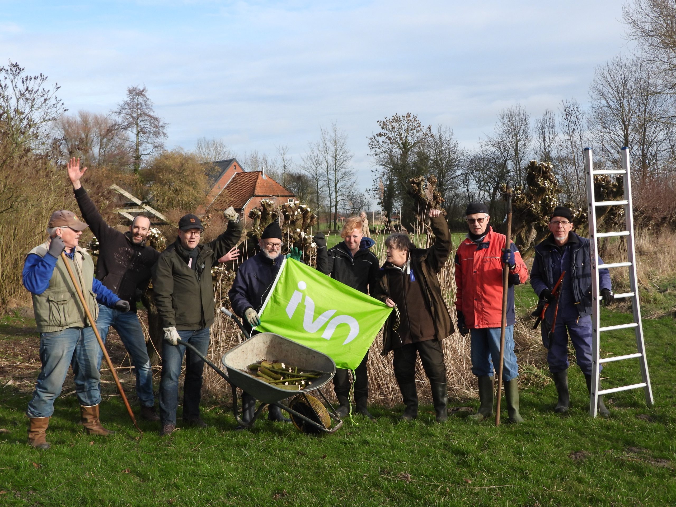 Groep mensen met tuingereedschap viert in natuurgebied naast een groene IVN-vlag en kruiwagen.
