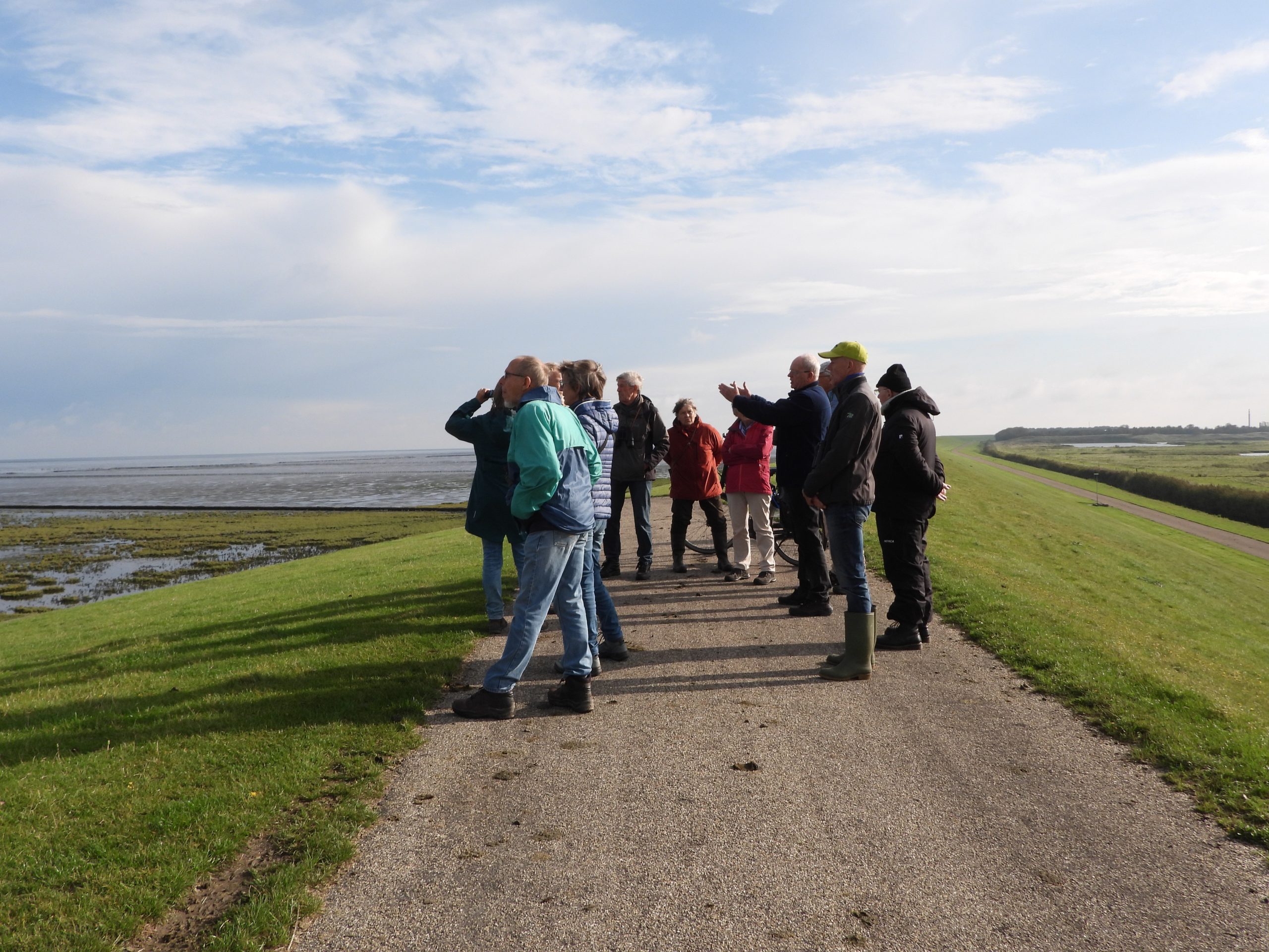 Groep mensen kijkt vanaf een dijk uit over een wijd, eb strand onder een deels bewolkte lucht.