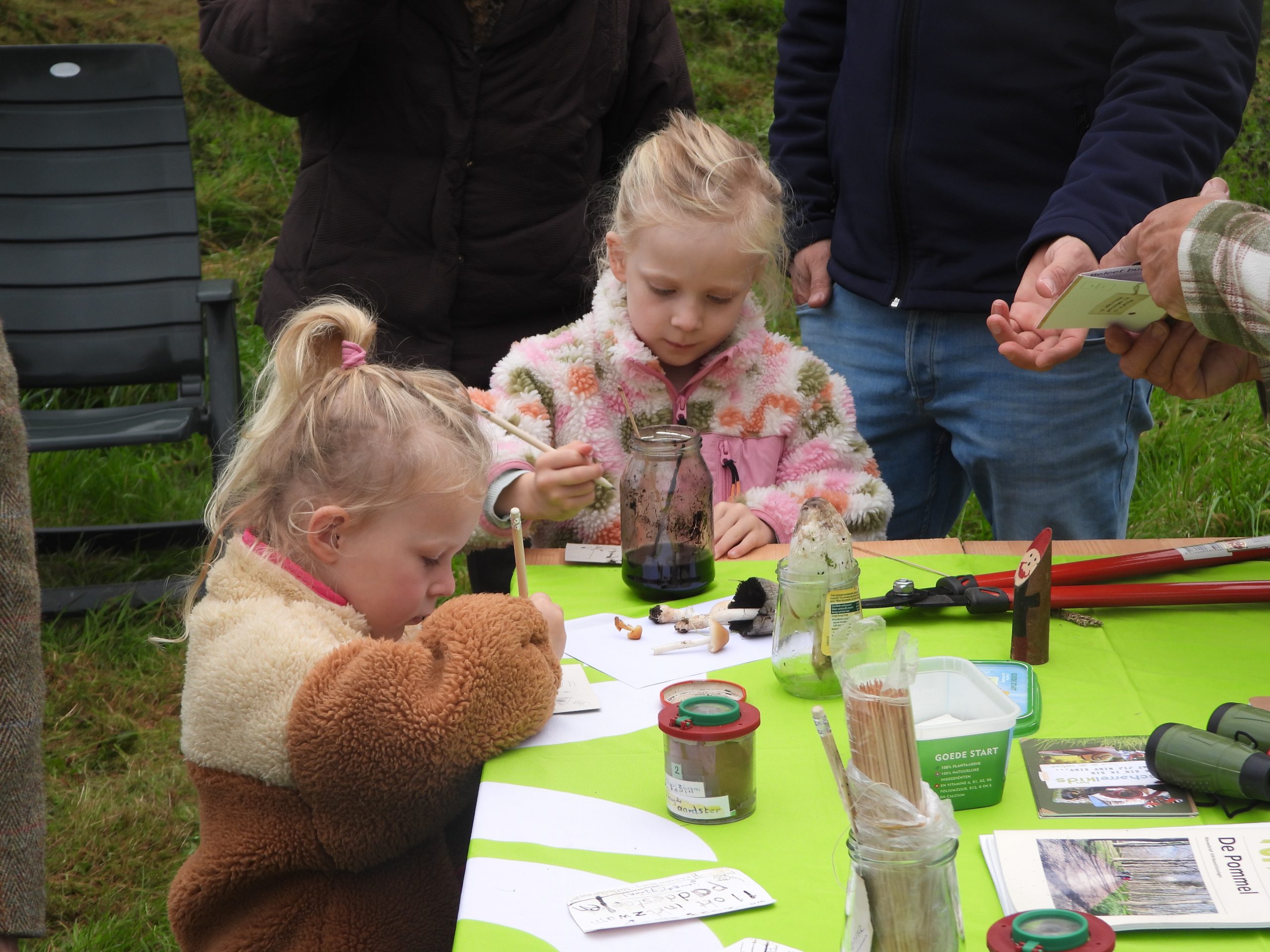 Twee kinderen knutselen aan tafel met potloden en potten op een buitenevenement.