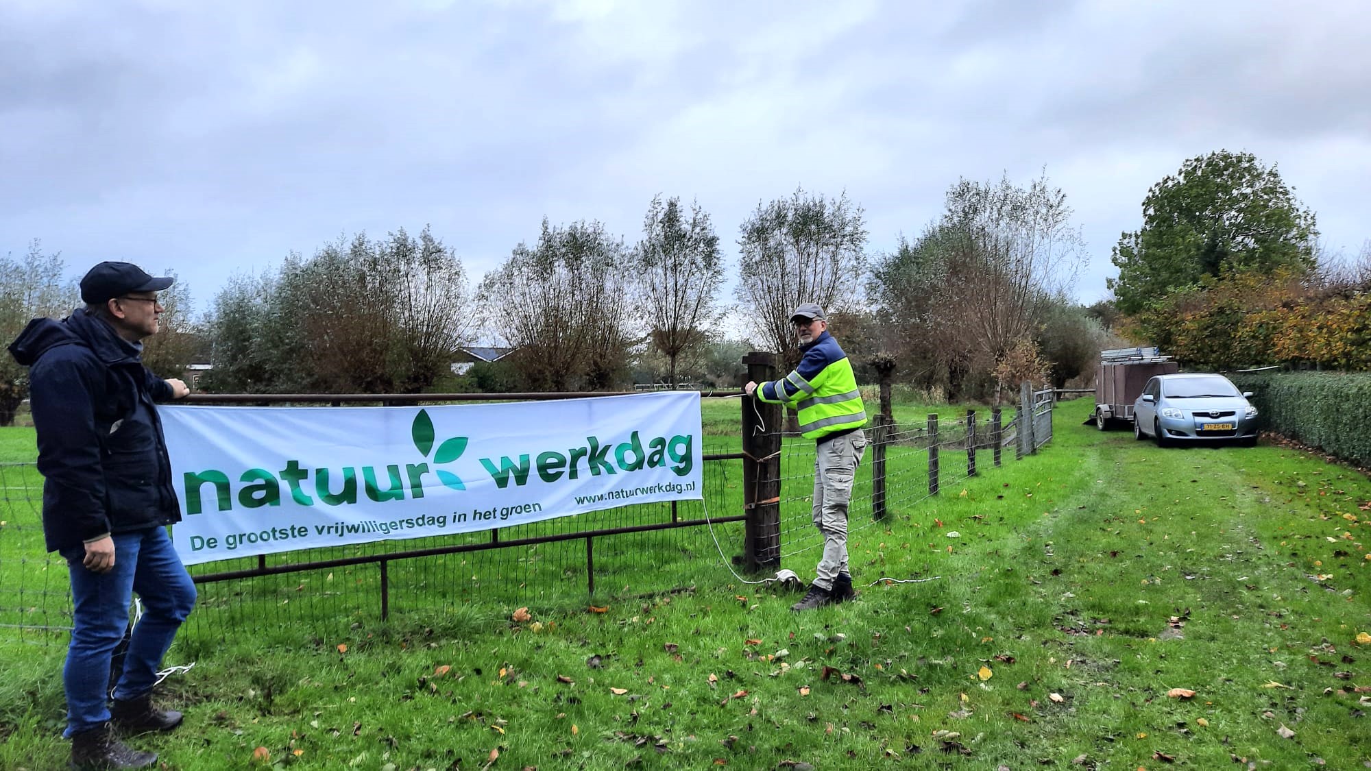 Twee mensen bevestigen een "Natuurwerkdag" spandoek aan een hek in een parksetting.