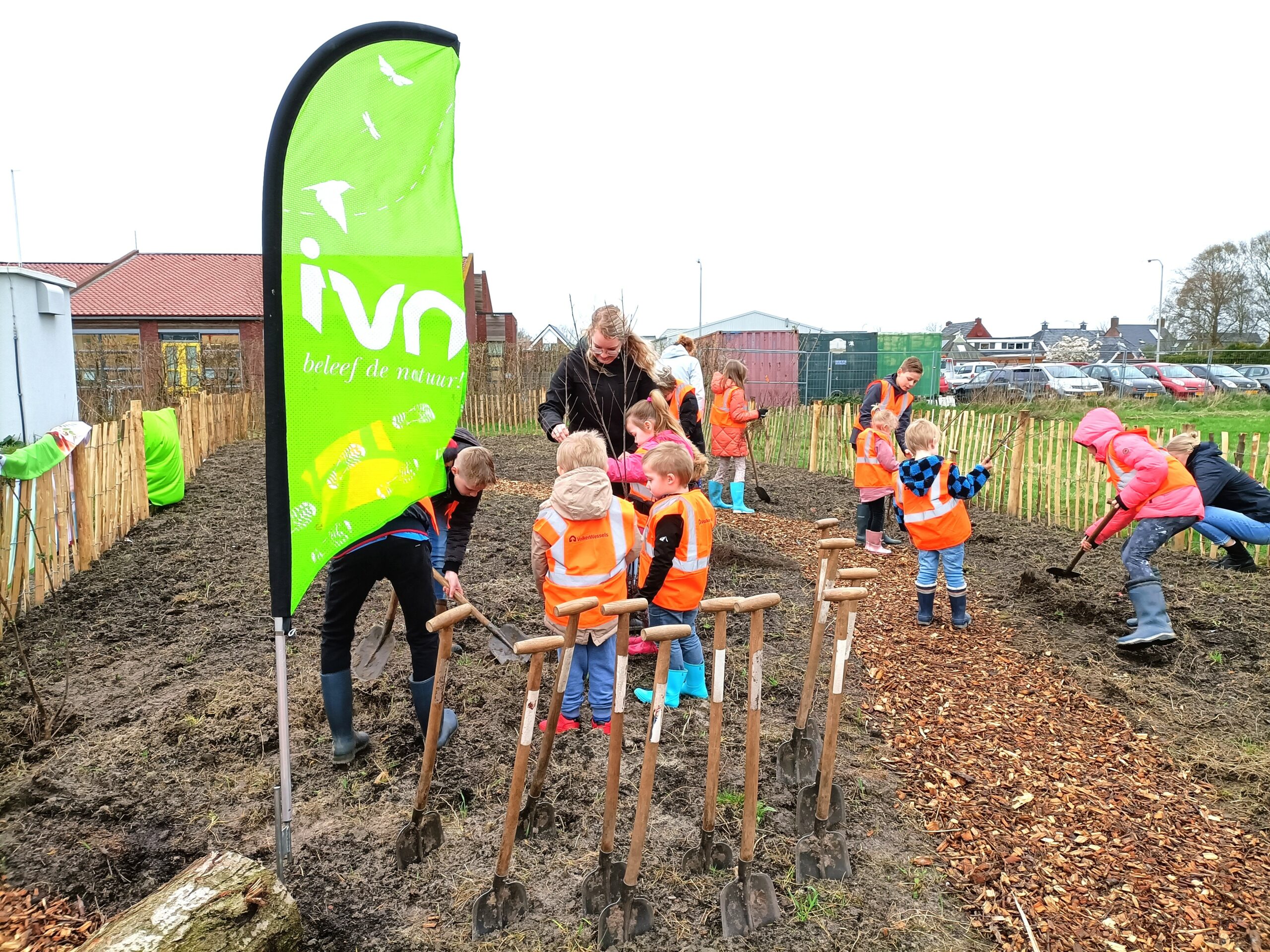 Kinderen in een tuin met oranje hesjes, graven onder begeleiding bij een groene IVN-vlag.
