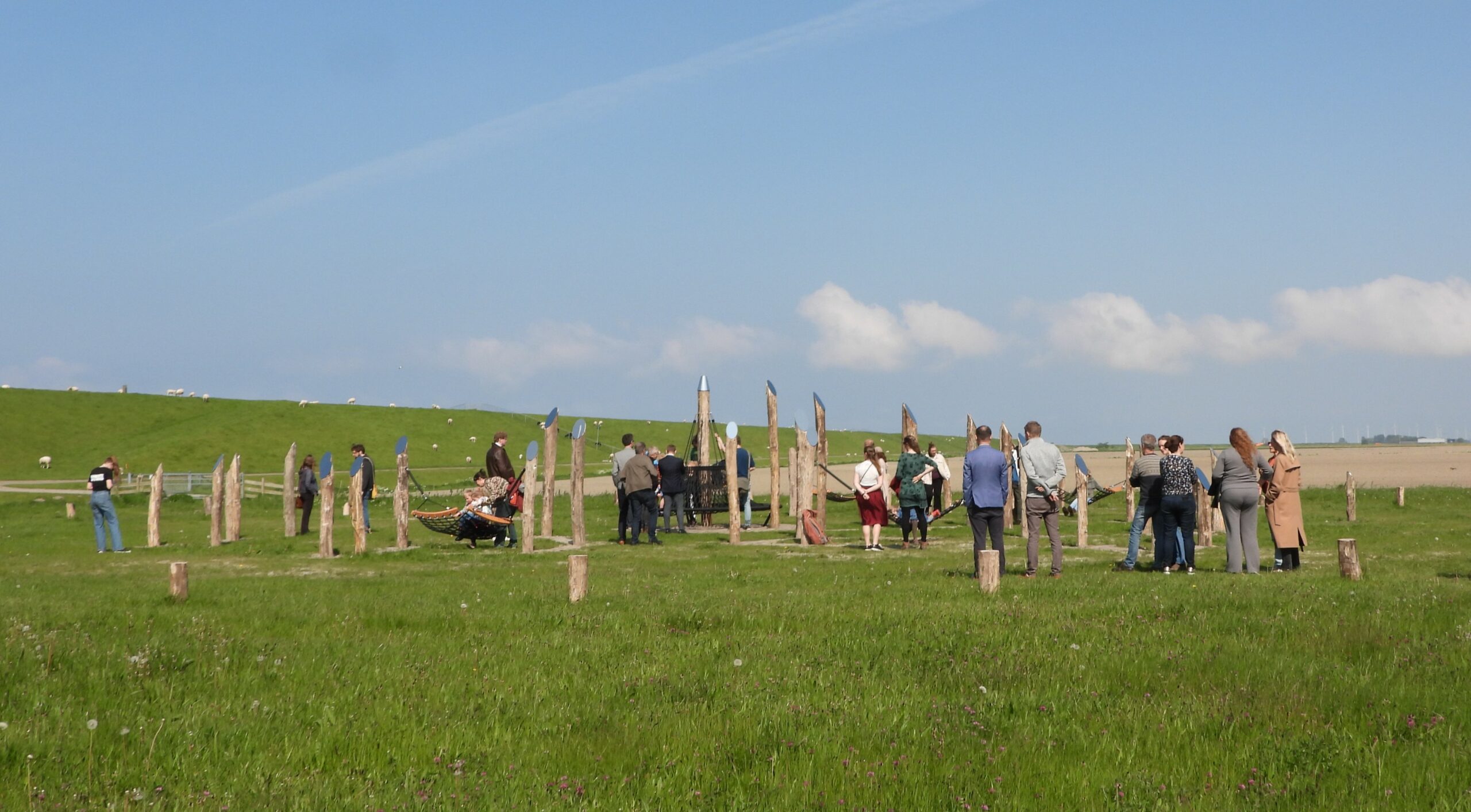 Groep mensen staat buiten in een grasveld, omgeven door houten palen, onder een blauwe hemel.