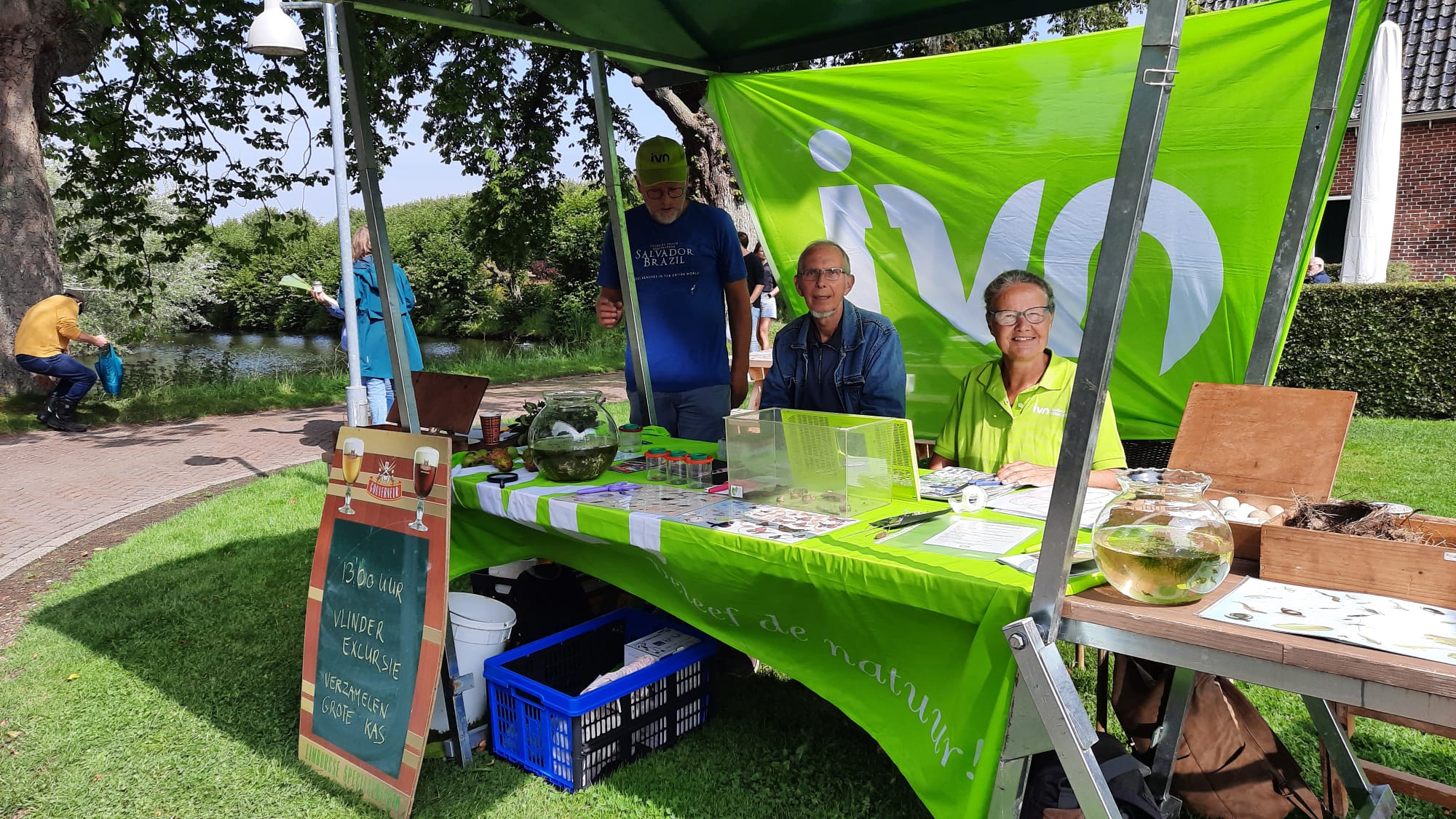 Drie mensen bij een informatiestand van IVN in een park met natuurproducten en folders.