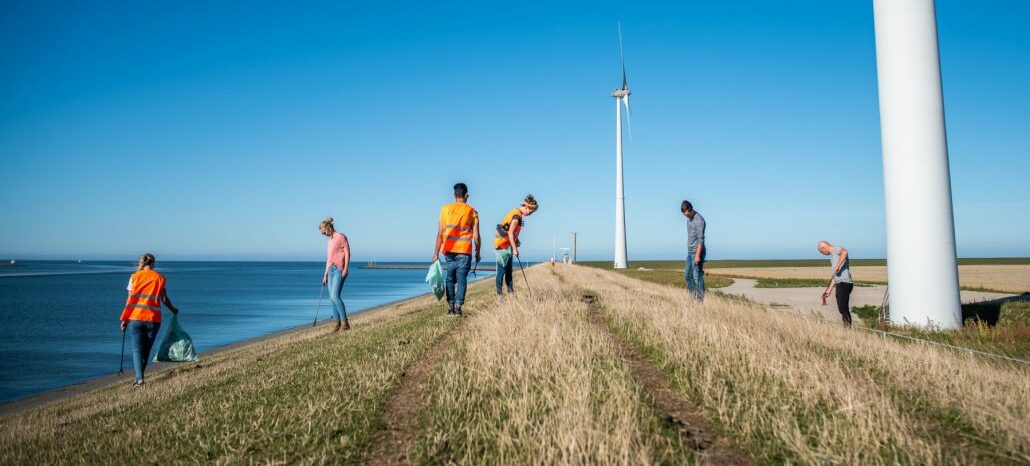 Mensen verzamelen afval langs een dijk bij windmolens en zee, onder een heldere blauwe lucht.
