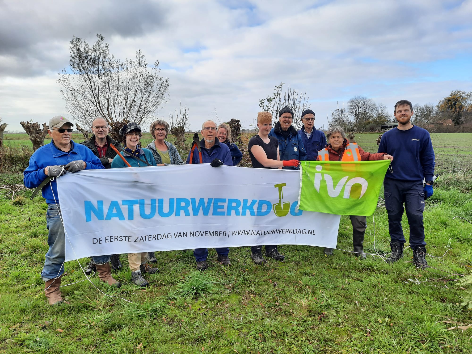 Groep vrijwilligers houdt spandoek met "Natuurwerkdag"; groene velden op de achtergrond.