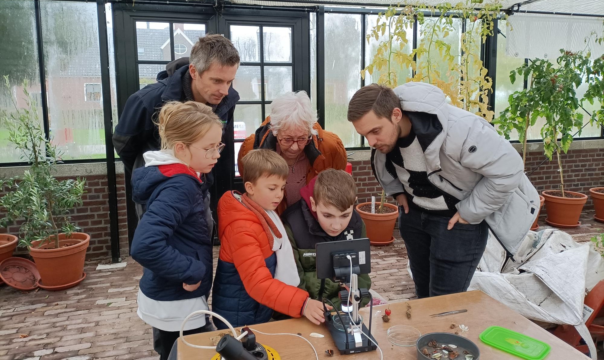 Een groep kinderen en volwassenen onderzoekt samen iets op een microscoop in een kas.