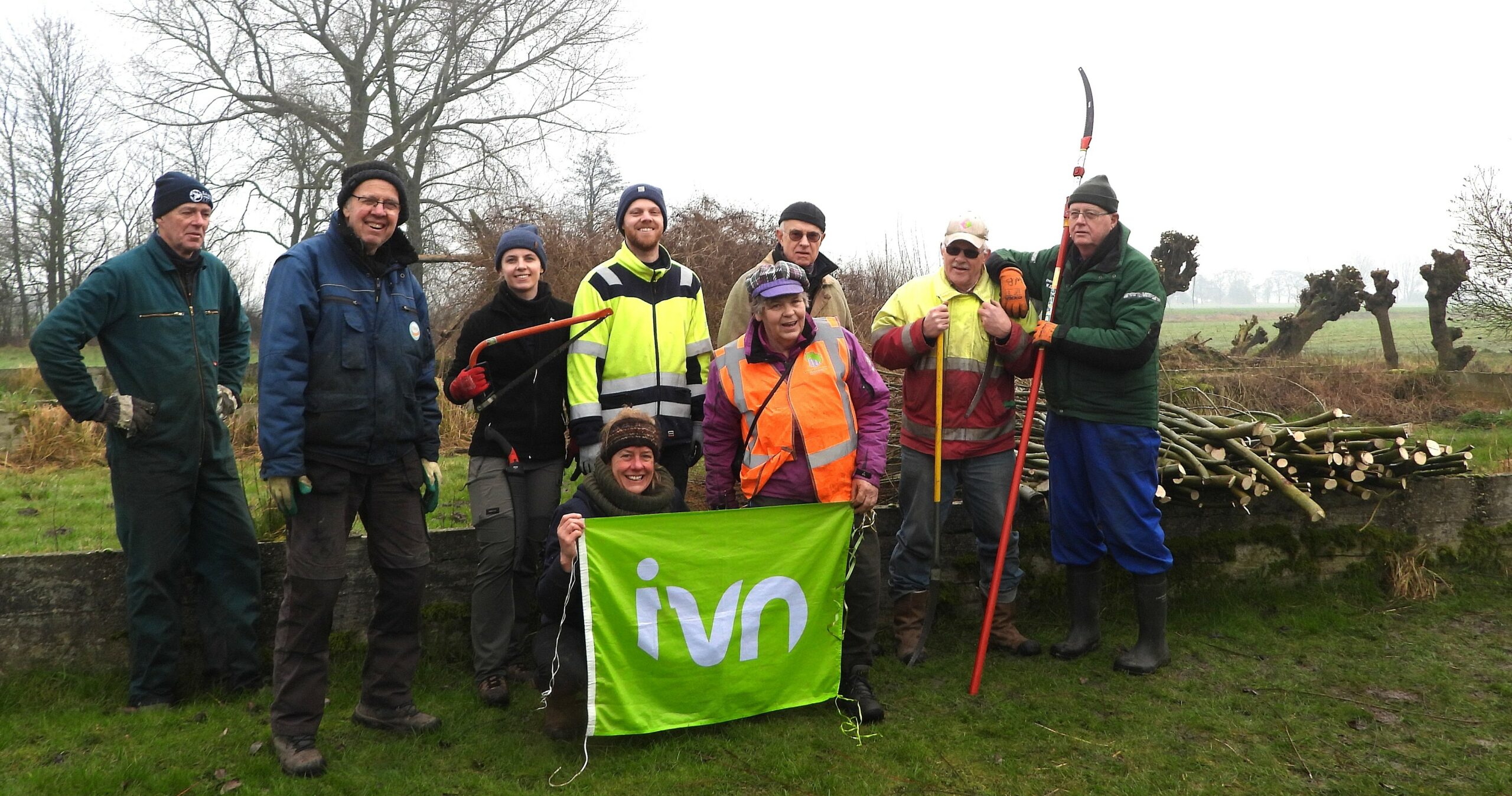 Groep vrijwilligers met gereedschap en IVN-banner in natuurgebied, bewolkte dag.