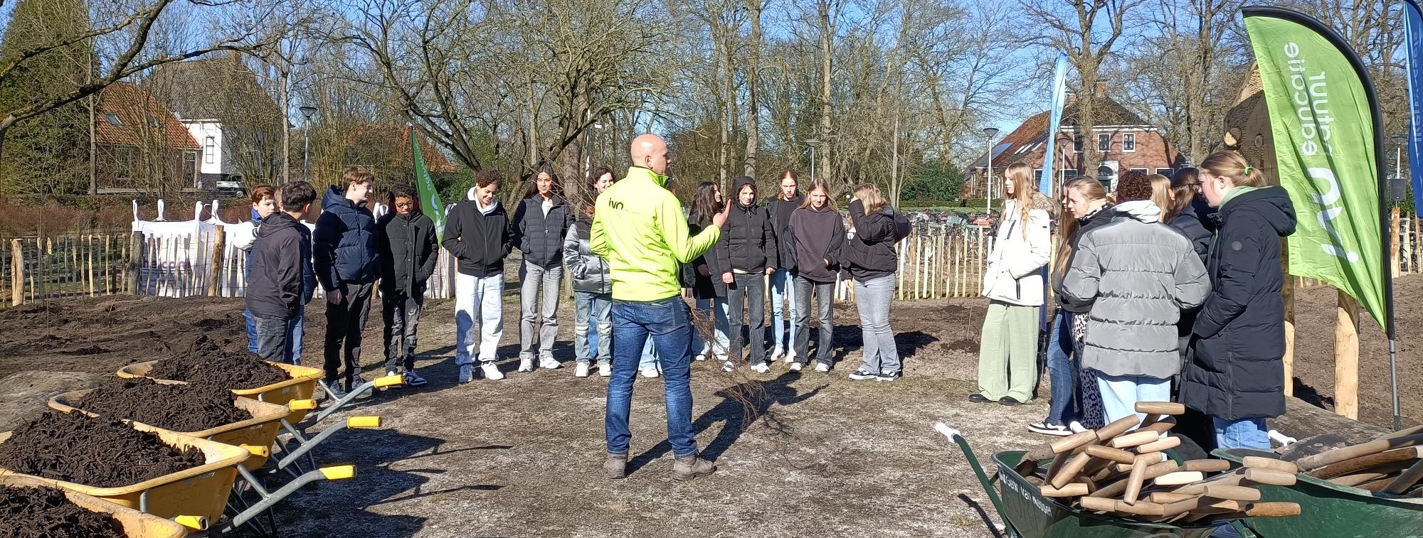 Een groep mensen verzameld buiten, klaar om te tuinieren met kruiwagens en gereedschap.