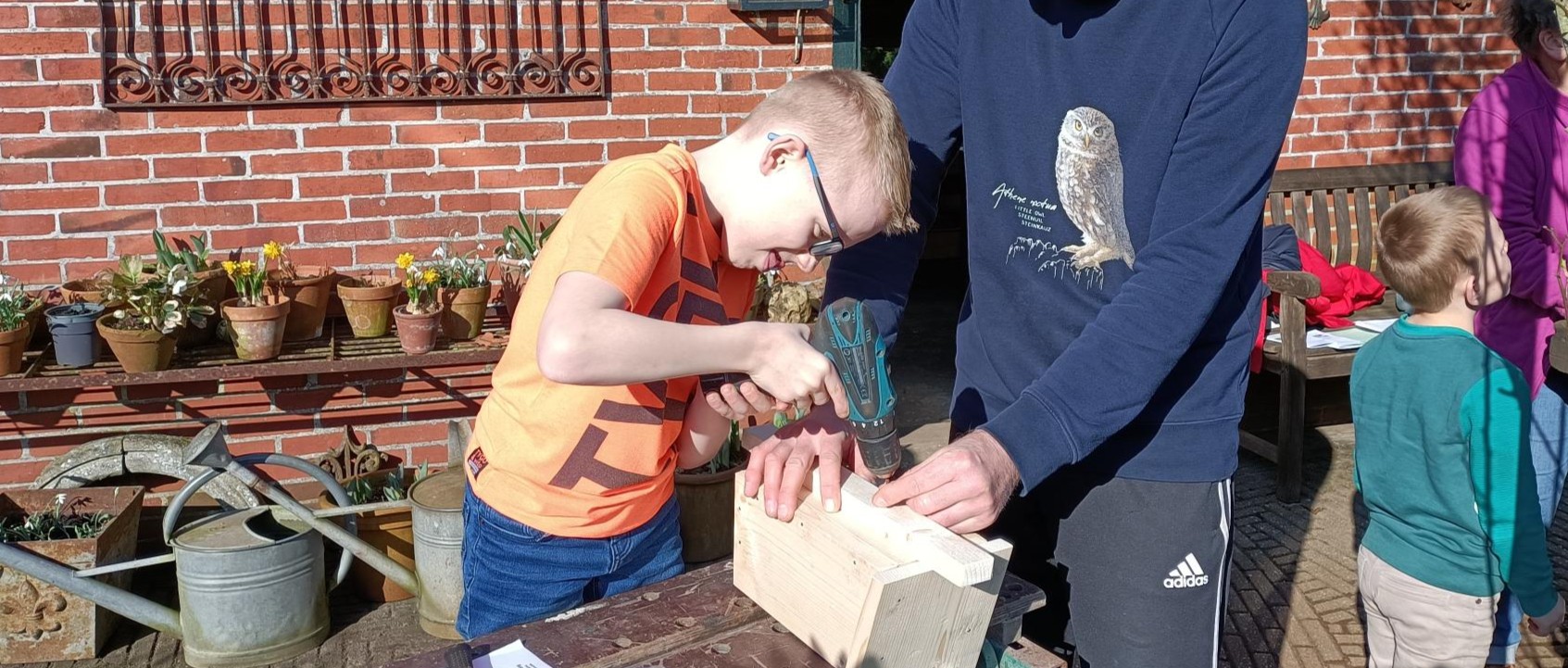 Een kind boort in hout met hulp van een volwassene buiten bij een bakstenen muur.