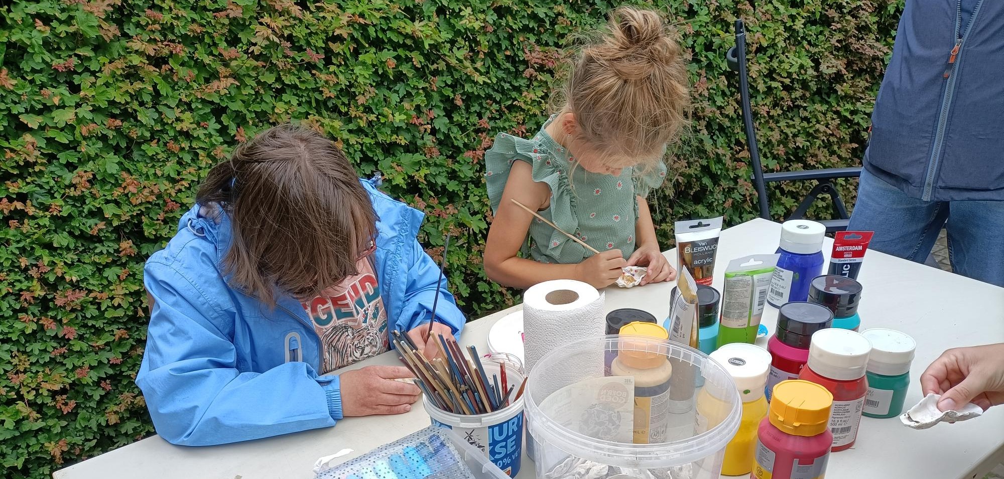 Twee kinderen schilderen kunstwerken aan een tafel buiten, omringd door verf en penselen.