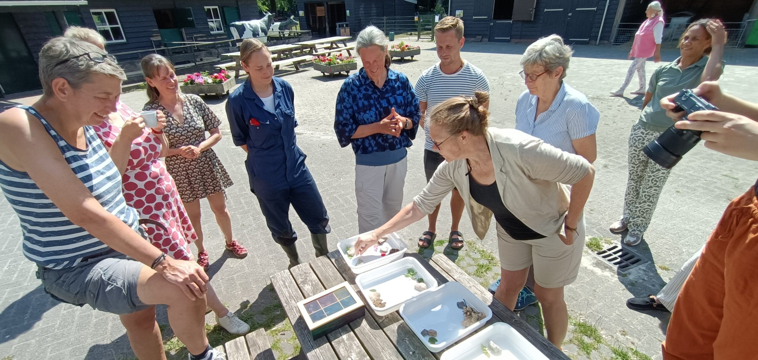 Groep mensen verzameld rond een picknicktafel met bakjes en objecten buiten.