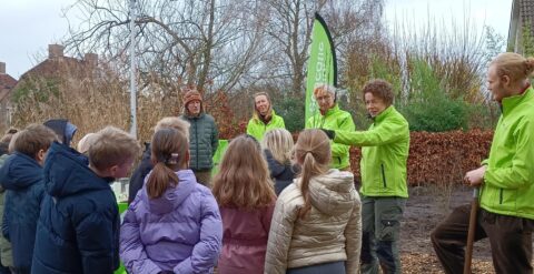 Groep volwassenen leert kinderen over de natuur buiten, met groene jassen en banner.