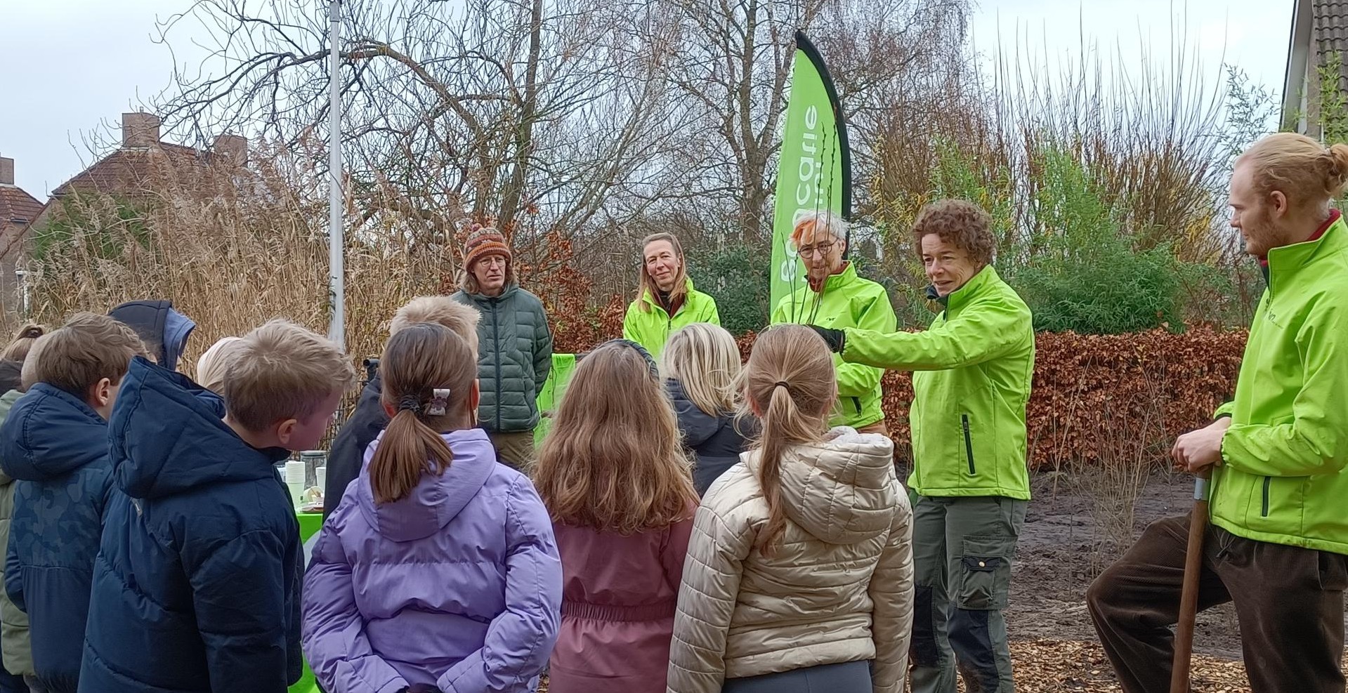 Groep volwassenen leert kinderen over de natuur buiten, met groene jassen en banner.