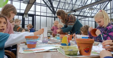Kinderen schilderen bloempotten in een serre, begeleid door een volwassene.
