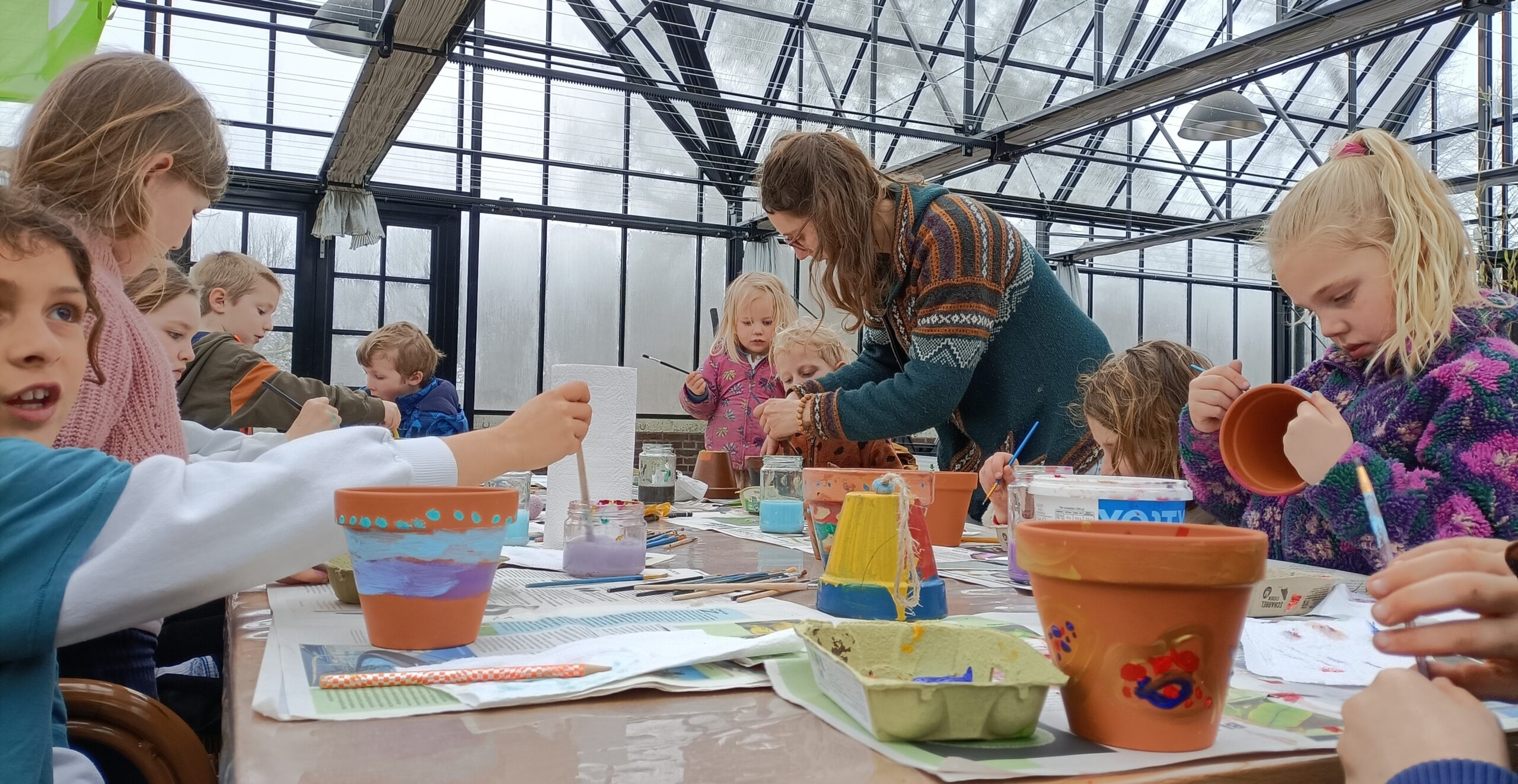 Kinderen schilderen bloempotten in een serre, begeleid door een volwassene.