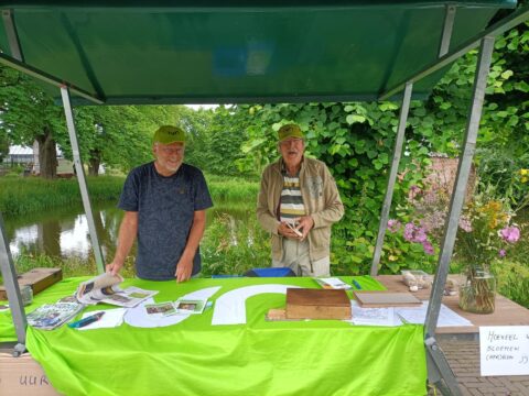 Twee mannen onder een groene kraam, omgeven door natuur, met papieren en bloemen op tafel.