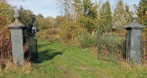 Open smeedijzeren poort tussen pilaren, leidt naar een groen, bosrijk pad.