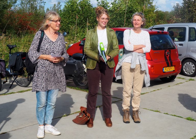 Drie vrouwen staan buiten naast auto's en fietsen bij een bijeenkomst in een zonnige omgeving.