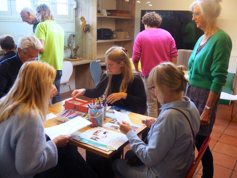 Groep mensen doet creatieve activiteiten rond een tafel met knutselmaterialen en kleurpotloden.