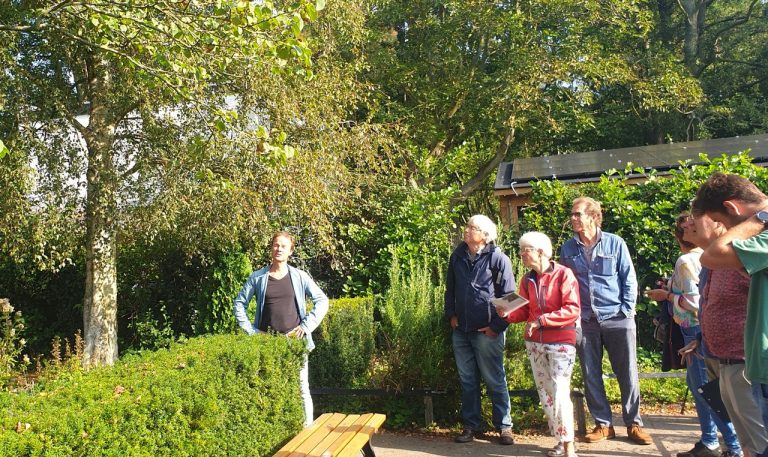 Mensen luisteren naar een spreker in een groene tuin onder een boom.