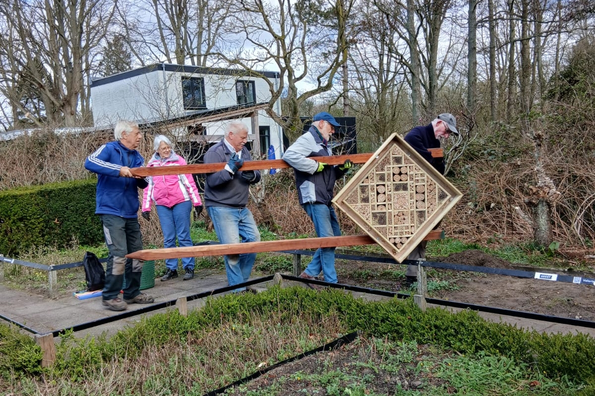 4 mensen die een groot en zwaar insectenhotel op palen dragen.