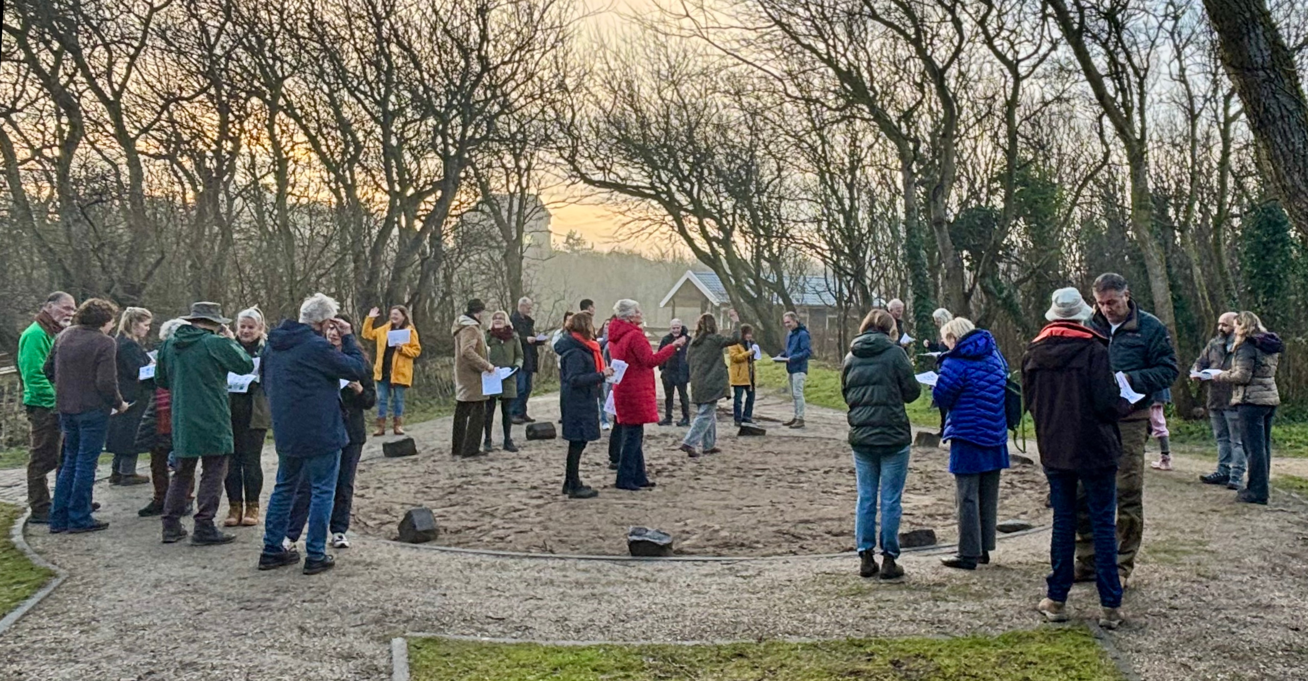 Een groep mensen staat in een kring buiten, omgeven door bomen bij zonsondergang.