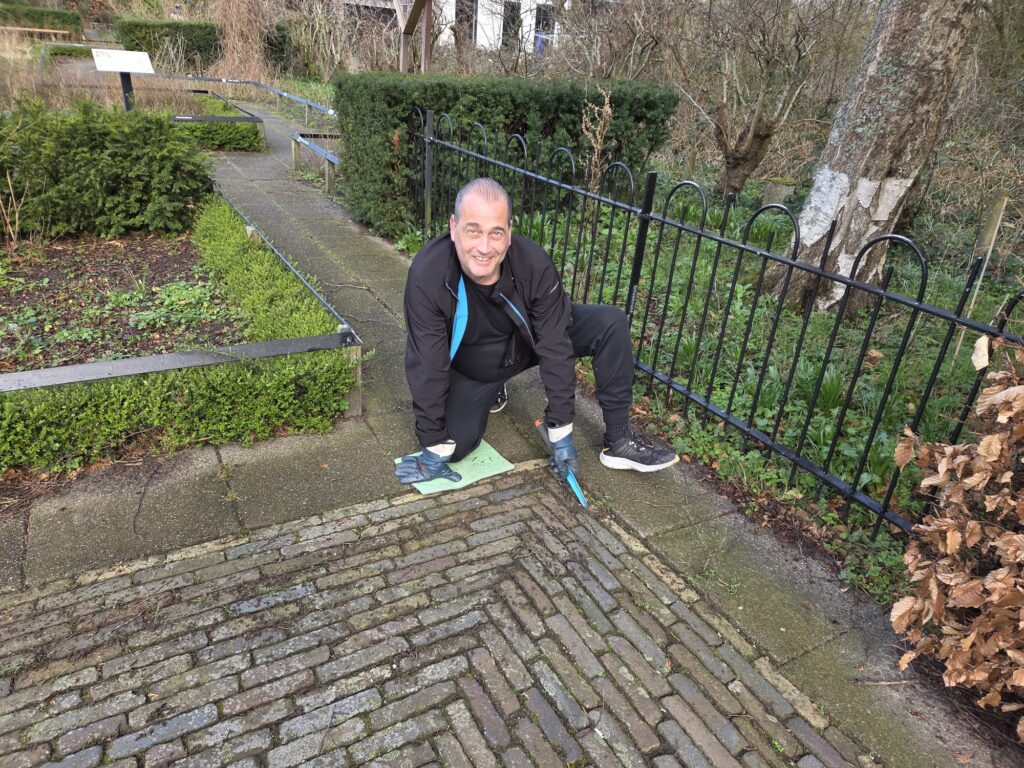 Man knielt op stoep met tuingereedschap, omringd door struiken en een boom.