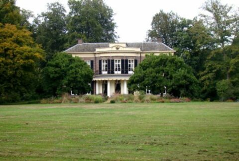 Een statig, klassiek herenhuis omgeven door bomen, met een grasveld op de voorgrond.