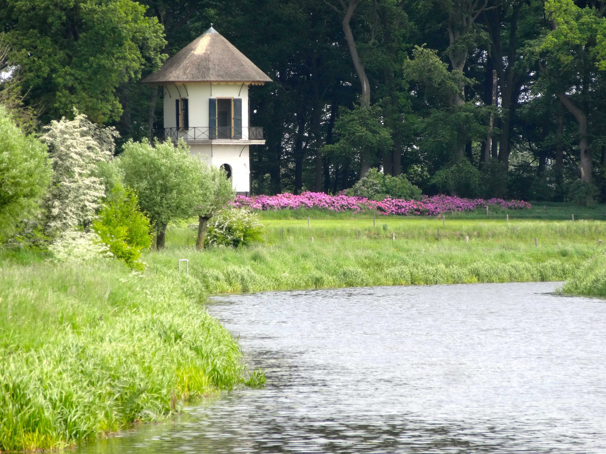 Toren met rieten dak naast een rivier, omgeven door groene bomen en roze bloemen in volle bloei.