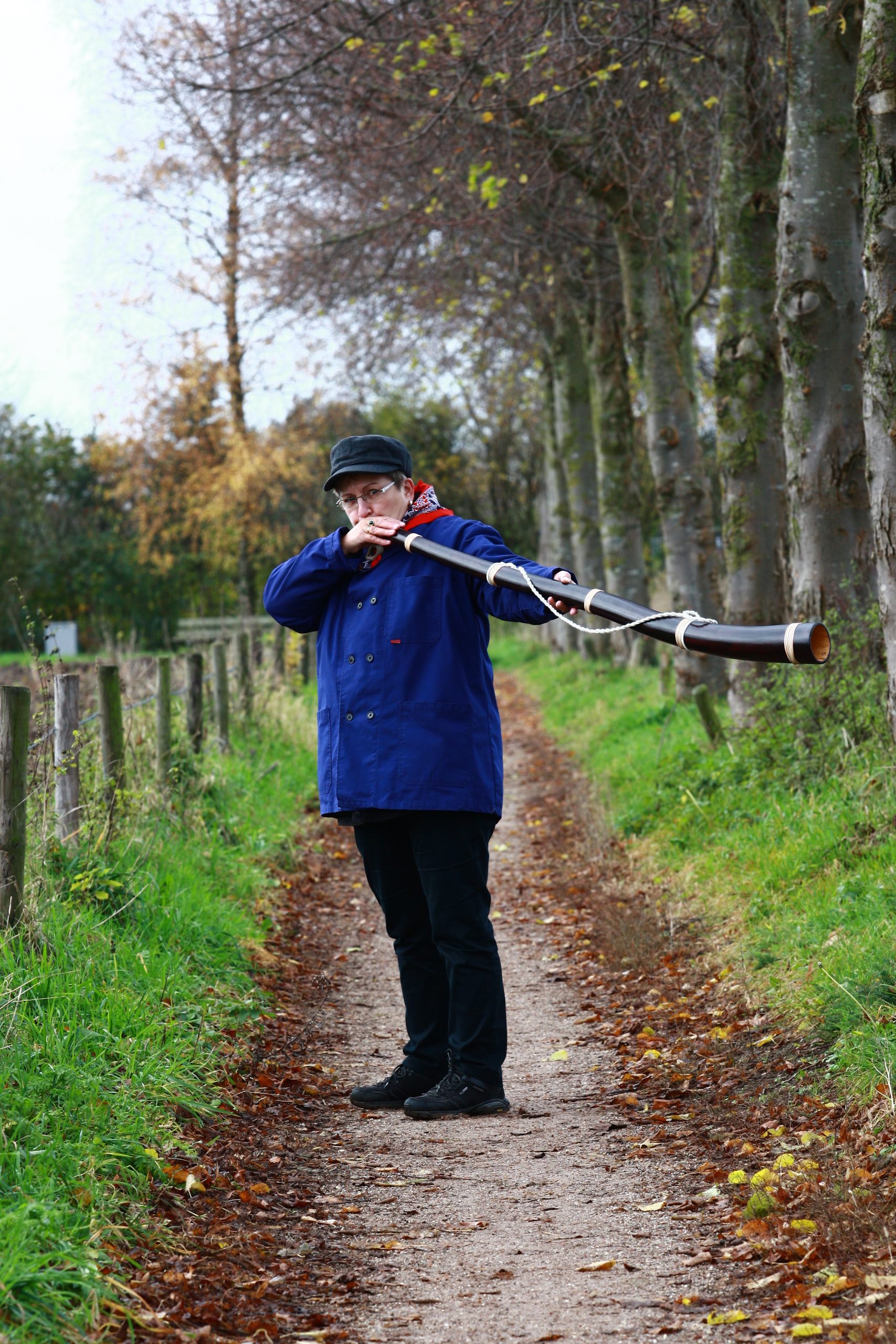 Persoon blaast didgeridoo op bospad in herfstomgeving.