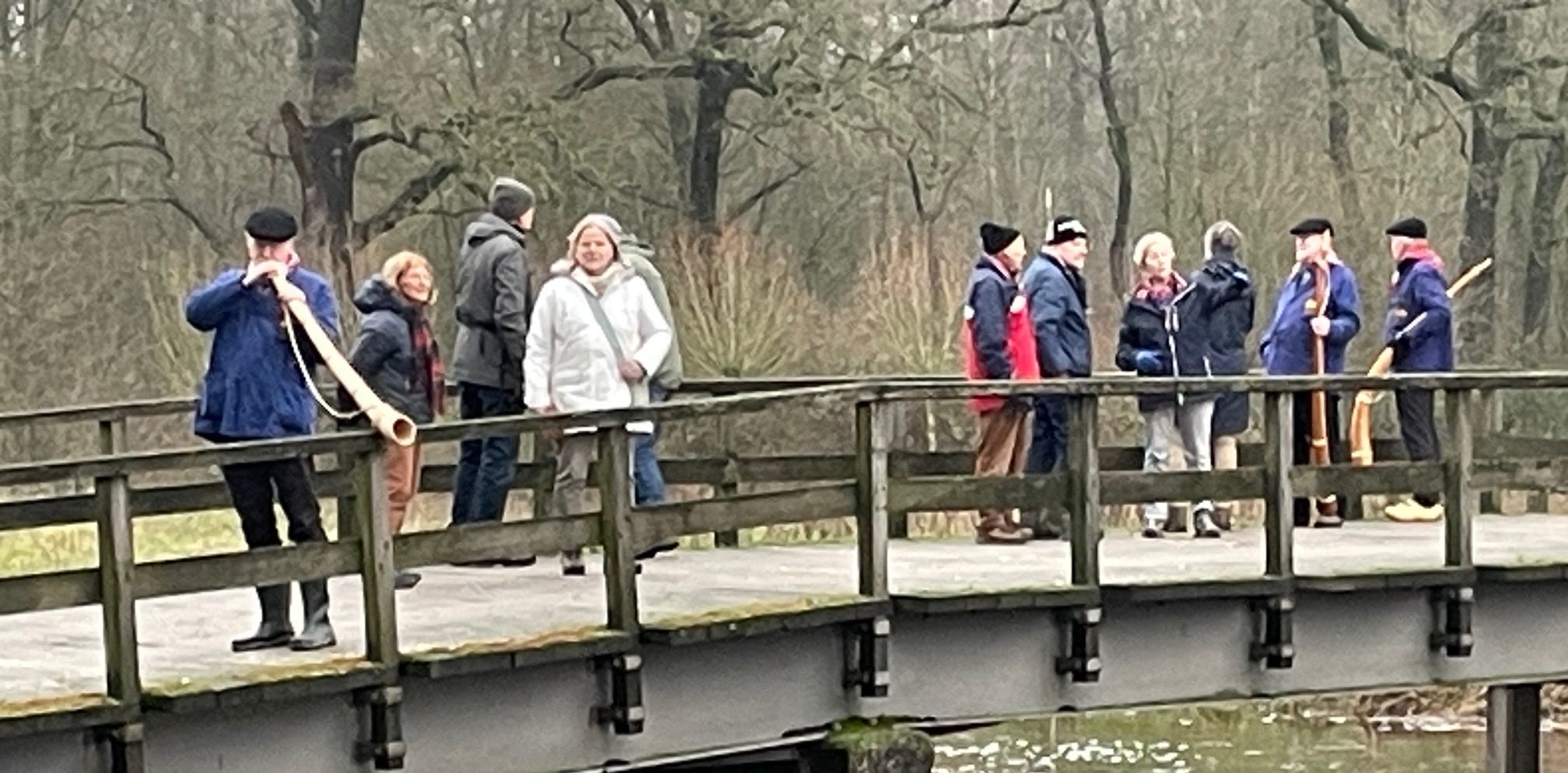 Mensen op een brug, één speelt een midwinterhoorn; winterse, bosrijke omgeving.