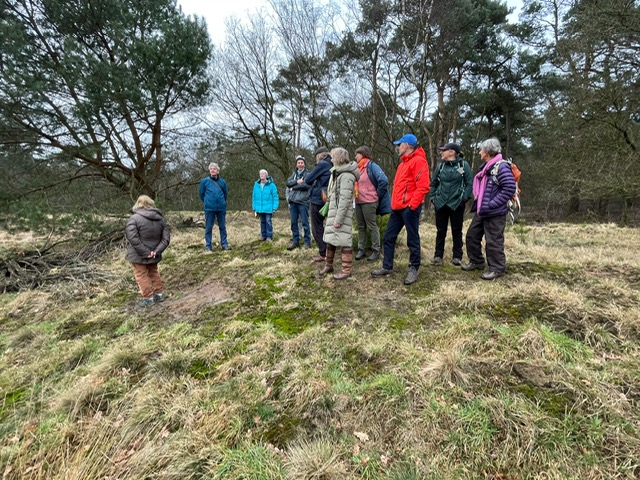 Een groep mensen luistert naar een spreker in een bosrijke omgeving met kale bomen.